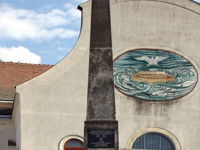 A tall stone obelisk war memorial stands in front of a modernist church facade featuring a large mosaic of a boat on waves and a cross at the top.