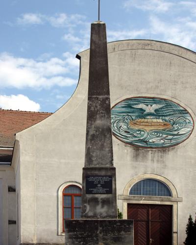 A tall stone obelisk war memorial stands in front of a modernist church facade featuring a large mosaic of a boat on waves and a cross at the top.