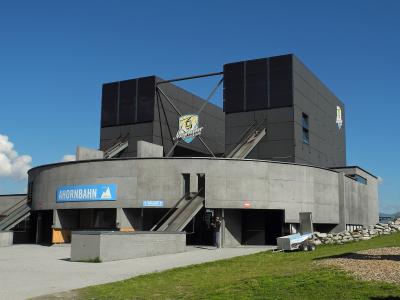 A modern concrete and dark-paneled cable car station building under a blue sky, featuring a prominent blue sign reading "AHORNBahn" and a logo for "Mayrhofner Bergbahnen" on the upper structure.