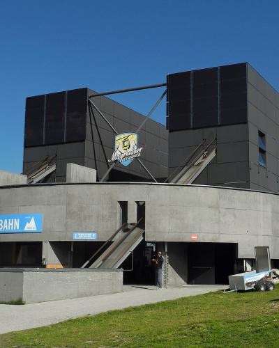 A modern concrete and dark-paneled cable car station building under a blue sky, featuring a prominent blue sign reading "AHORNBahn" and a logo for "Mayrhofner Bergbahnen" on the upper structure.