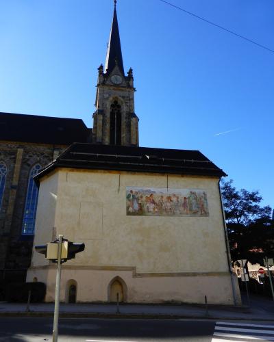 A low-angle view of a church with a tall, dark spire and a clock tower, featuring a large mural on the side of an attached lower structure, with a traffic light in the foreground.