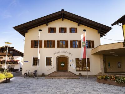 The image shows a cream-colored municipal building with brown shutters, a large wooden door, and the word "GEMEINDEAMT" (Town Hall) arched above the entrance, flanked by an Austrian flag and a regional flag.
