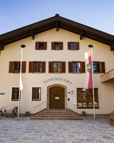 The image shows a cream-colored municipal building with brown shutters, a large wooden door, and the word "GEMEINDEAMT" (Town Hall) arched above the entrance, flanked by an Austrian flag and a regional flag.
