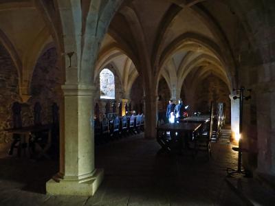 The image shows the interior of a stone hall with ribbed vaulted ceilings, long wooden tables, and benches, where two people are standing in the background.