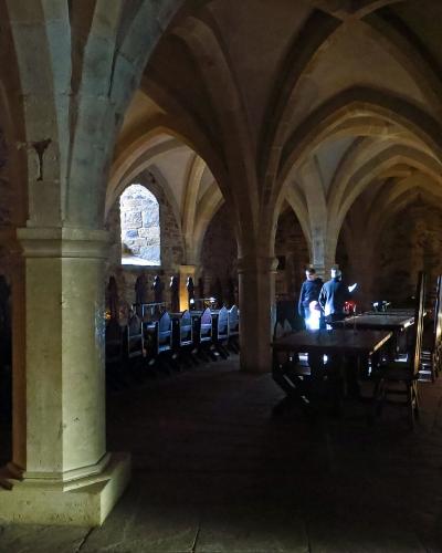 The image shows the interior of a stone hall with ribbed vaulted ceilings, long wooden tables, and benches, where two people are standing in the background.