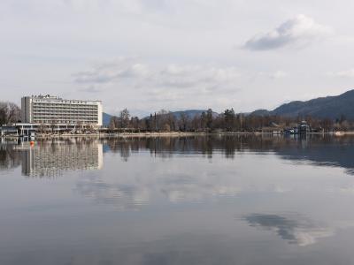 A large white hotel building with "PARKHOTEL" visible on the roof, situated on the shore of a calm lake with mountains in the background.