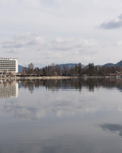 A large white hotel building with "PARKHOTEL" visible on the roof, situated on the shore of a calm lake with mountains in the background.