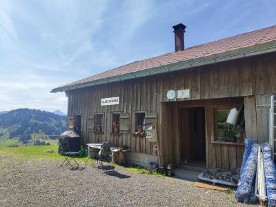 A wooden alpine hut with a red tiled roof, featuring a sign reading "ALPE ROHNE" on the facade, set against a backdrop of green hills and blue sky.