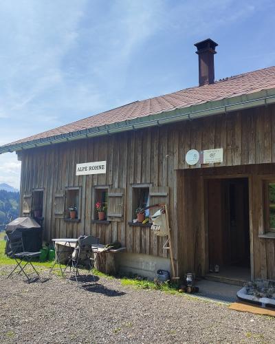 A wooden alpine hut with a red tiled roof, featuring a sign reading "ALPE ROHNE" on the facade, set against a backdrop of green hills and blue sky.