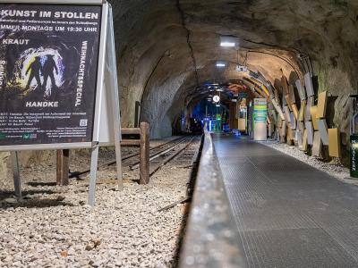An underground tunnel station with a railway track, a platform, and a large poster advertising "KUNST IM STOLLEN" (Art in the Tunnel) at the Schloßbergs.