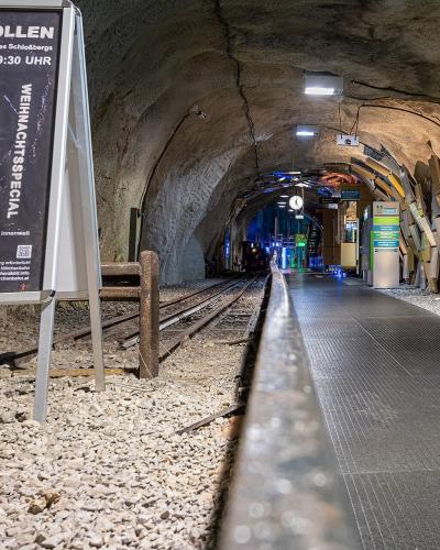 An underground tunnel station with a railway track, a platform, and a large poster advertising "KUNST IM STOLLEN" (Art in the Tunnel) at the Schloßbergs.