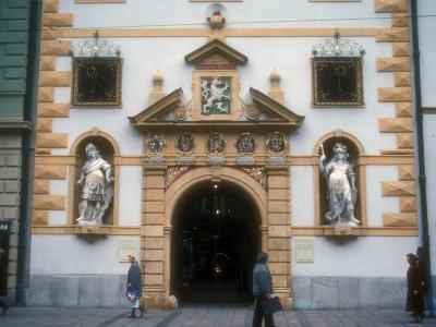 A street-level view of the ornate Renaissance entrance facade of a large building, featuring a central archway flanked by two statues in niches, a coat of arms above the arch, and rusticated stone quoins on the corners.