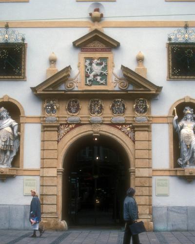 A street-level view of the ornate Renaissance entrance facade of a large building, featuring a central archway flanked by two statues in niches, a coat of arms above the arch, and rusticated stone quoins on the corners.