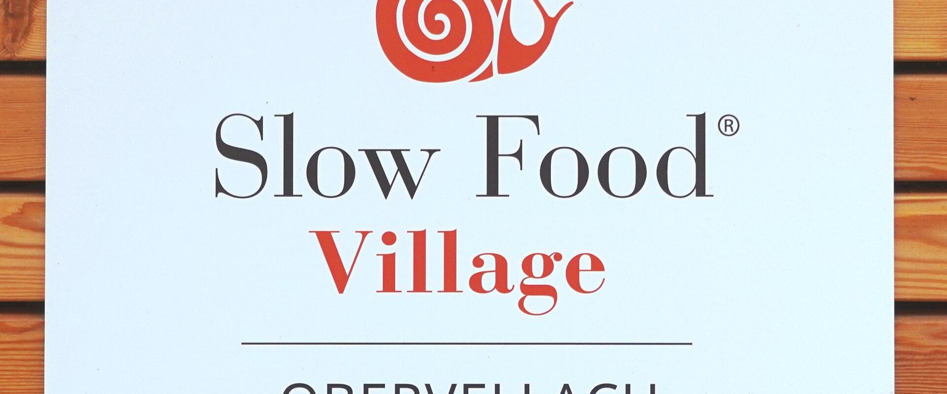 A white sign mounted on a wooden slat wall, displaying the Slow Food Village logo and text "Ein Ort guten Lebens", "Slow Food® Village", "OBERVELLACH KÄRNTEN", and logos for Slow Food Kärnten, LGO, and Land Kärnten.