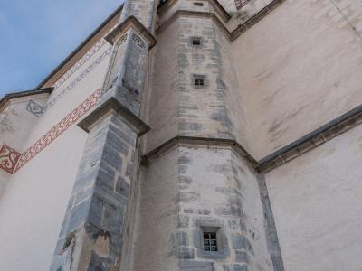A low-angle view of a stone church tower and adjacent wall featuring decorative frescoes, taken under a blue sky.