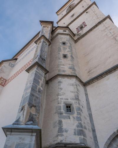 A low-angle view of a stone church tower and adjacent wall featuring decorative frescoes, taken under a blue sky.