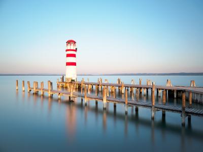 A red and white striped lighthouse standing at the end of a long wooden pier extending into calm water, with distant hills on the horizon.