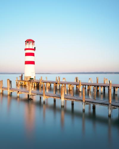 A red and white striped lighthouse standing at the end of a long wooden pier extending into calm water, with distant hills on the horizon.