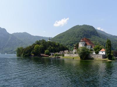 A view of the pilgrimage island of Mariazell on Traunsee, featuring the white church with its distinctive onion-domed towers and surrounding buildings, set against the backdrop of the Traunstein mountain.