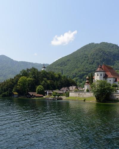 A view of the pilgrimage island of Mariazell on Traunsee, featuring the white church with its distinctive onion-domed towers and surrounding buildings, set against the backdrop of the Traunstein mountain.