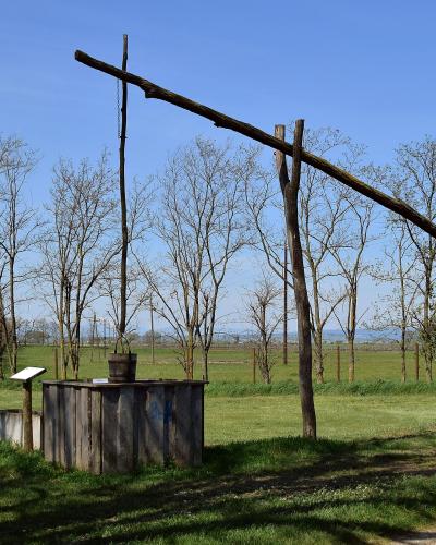 A wooden well structure with a long horizontal beam and bucket, situated in a grassy field with bare trees and a dirt path under a blue sky.
