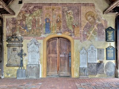 A covered porch or loggia featuring a central wooden double door, flanked by numerous stone grave markers and memorial plaques, with a large, faded fresco depicting the Crucifixion and saints on the wall above.