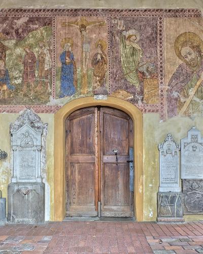 A covered porch or loggia featuring a central wooden double door, flanked by numerous stone grave markers and memorial plaques, with a large, faded fresco depicting the Crucifixion and saints on the wall above.