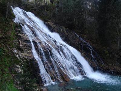 A wide waterfall cascading down a steep, layered rock face into a pool, surrounded by a forest with some snow visible on the banks.