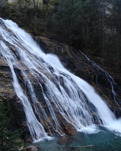 A wide waterfall cascading down a steep, layered rock face into a pool, surrounded by a forest with some snow visible on the banks.
