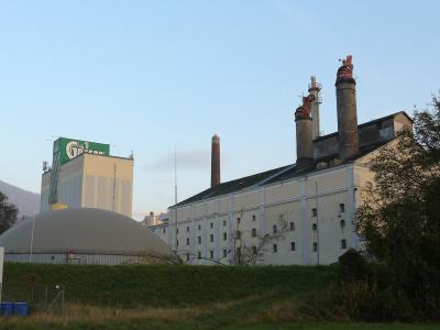 An industrial brewery complex featuring a large white building with multiple chimneys, a biogas digester dome, and a tall silo structure with a green sign displaying the "Gösser" logo.