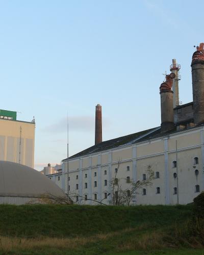 An industrial brewery complex featuring a large white building with multiple chimneys, a biogas digester dome, and a tall silo structure with a green sign displaying the "Gösser" logo.
