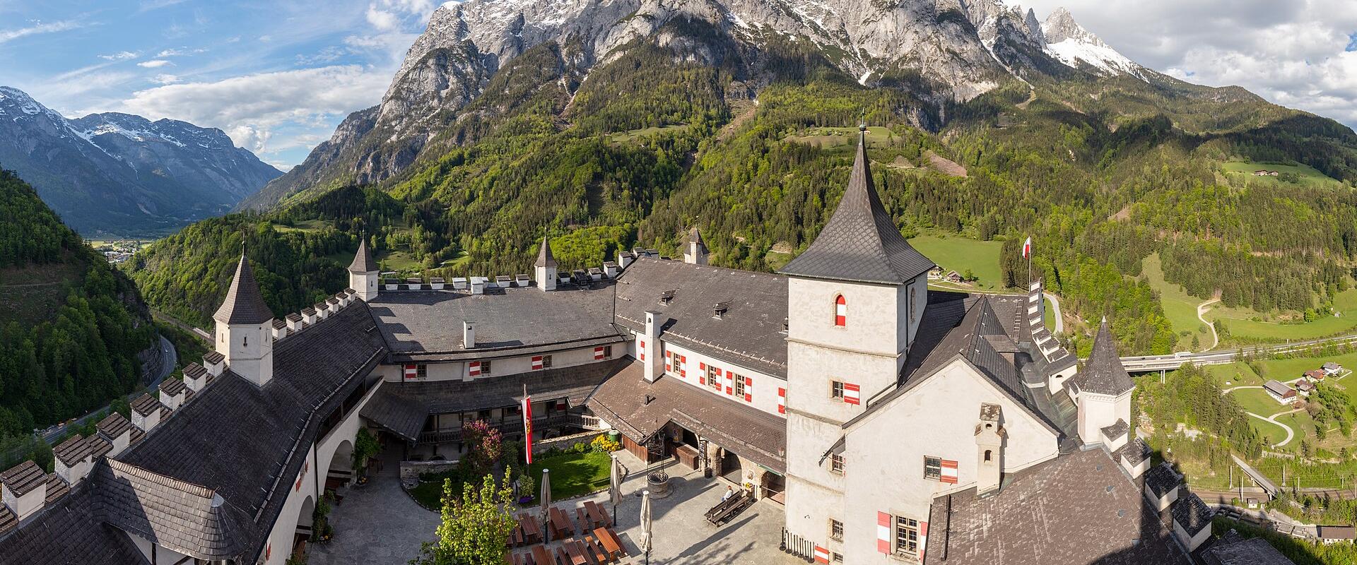 A high-angle view of a medieval castle courtyard with white walls, dark slate roofs, pointed towers, and red-and-white Austrian flags, set against a backdrop of steep green hills and snow-capped mountains under a blue sky with scattered clouds.