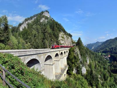 A red passenger train crosses a long, multi-arched stone viaduct built into a steep, forested mountainside under a blue sky.