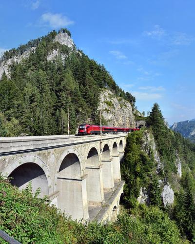 A red passenger train crosses a long, multi-arched stone viaduct built into a steep, forested mountainside under a blue sky.