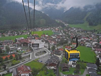 A yellow cable car gondola with "PENKENBAHN" written on its side is suspended on cables above a town in a green valley, with a station building visible below and mountains in the background.