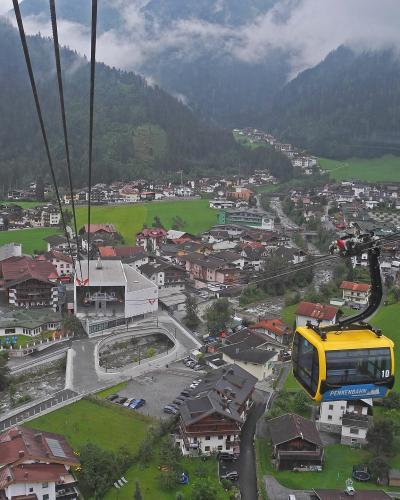 A yellow cable car gondola with "PENKENBAHN" written on its side is suspended on cables above a town in a green valley, with a station building visible below and mountains in the background.