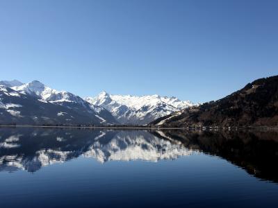 A panoramic view of a calm alpine lake reflecting snow-capped mountains under a clear blue sky, with a forested slope on the right and distant buildings along the far shore.