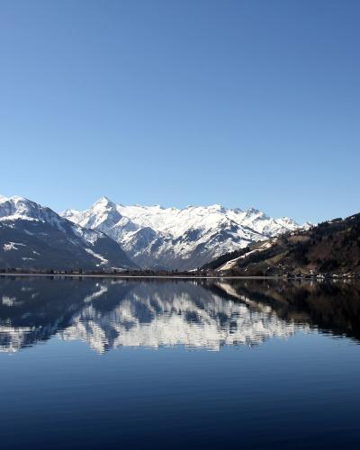 A panoramic view of a calm alpine lake reflecting snow-capped mountains under a clear blue sky, with a forested slope on the right and distant buildings along the far shore.