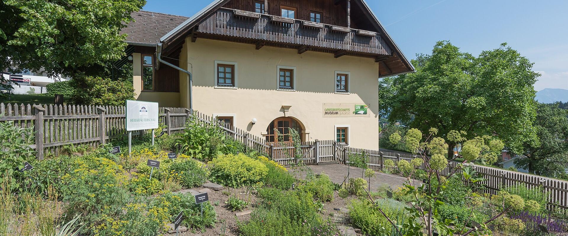 A traditional yellow building with a wooden balcony and dark roof, surrounded by a herb garden with labeled plants, a wooden fence, and a sign reading "Landwirtschaftsmuseum Schloss Ehrenthal" on the wall; another sign near the fence says "Herbari