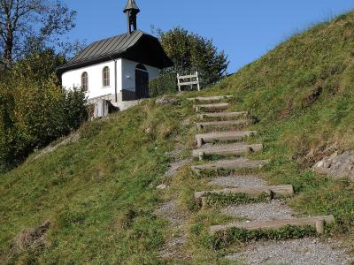 A small white chapel with a dark roof and steeple sits atop a grassy hill, accessed by a gravel path with wooden steps.