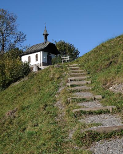 A small white chapel with a dark roof and steeple sits atop a grassy hill, accessed by a gravel path with wooden steps.