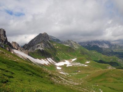 A mountain landscape showing green alpine slopes with patches of snow and rocky peaks under a cloudy sky.