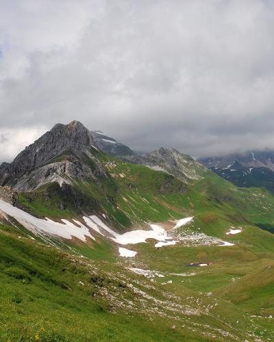 A mountain landscape showing green alpine slopes with patches of snow and rocky peaks under a cloudy sky.