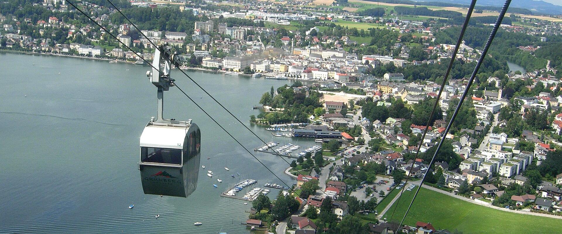 A cable car cabin suspended over a lake, with a town and hills in the background; the cabin bears the logo "Gmundenbahn" and is positioned above a marina and residential area.