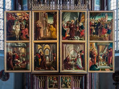 A large, multi-panelled Gothic altarpiece with numerous painted scenes, displayed inside a church with stained-glass windows.
