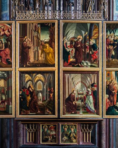 A large, multi-panelled Gothic altarpiece with numerous painted scenes, displayed inside a church with stained-glass windows.
