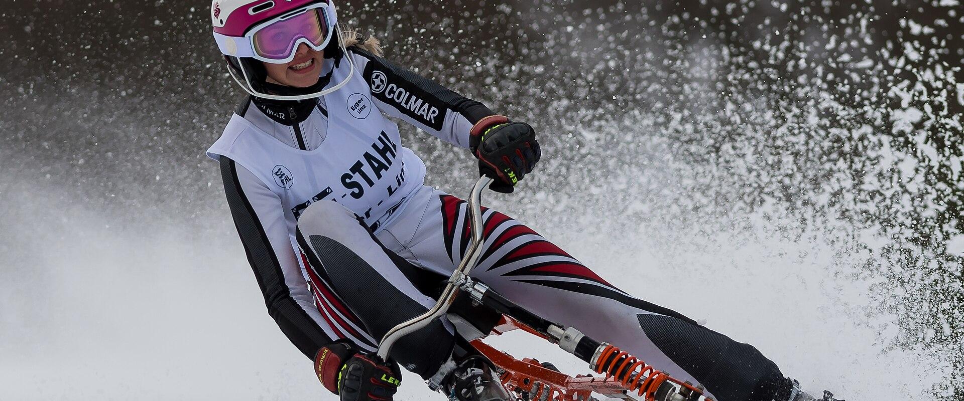A female athlete in a white, black, and red racing suit and helmet is riding a skibob (a seated ski vehicle with handlebars) down a snowy slope, kicking up snow spray.