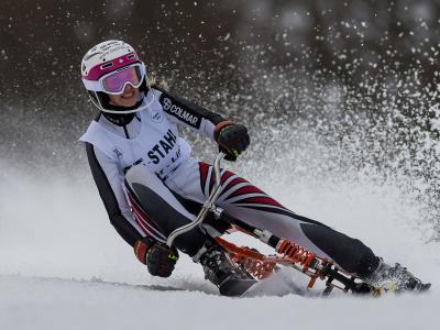 A female athlete in a white, black, and red racing suit and helmet is riding a skibob (a seated ski vehicle with handlebars) down a snowy slope, kicking up snow spray.
