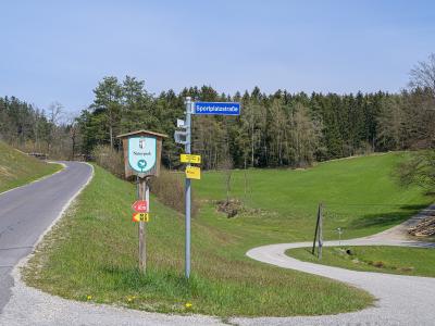 A roadside scene in a rural, hilly area with green fields and a forest background, featuring a wooden signpost with a "Naturpark" logo, a blue street sign reading "Sportplatzstraße", and yellow hiking trail markers.
