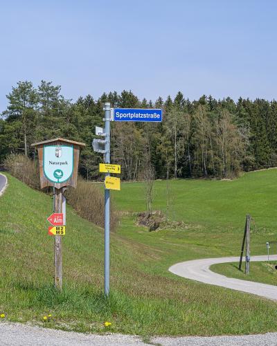 A roadside scene in a rural, hilly area with green fields and a forest background, featuring a wooden signpost with a "Naturpark" logo, a blue street sign reading "Sportplatzstraße", and yellow hiking trail markers.